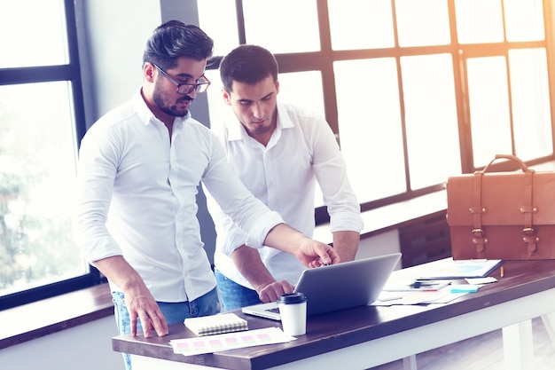 Due giovani imprenditori che lavorano al computer portatile in ufficio moderno. Effetto pellicola, riflesso lente