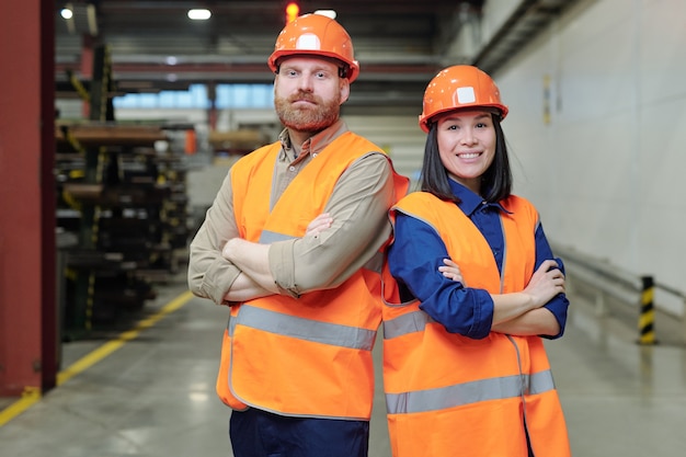 Due giovani ingegneri di successo in elmetto protettivo e abbigliamento da lavoro che incrociano le braccia per il petto mentre si trovano in fabbrica