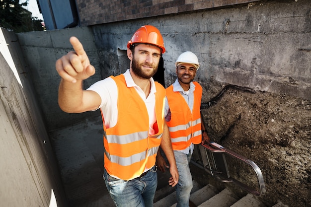 Due giovani ingegneri maschi in uniforme e elmetti protettivi che lavorano in cantiere, primi piani