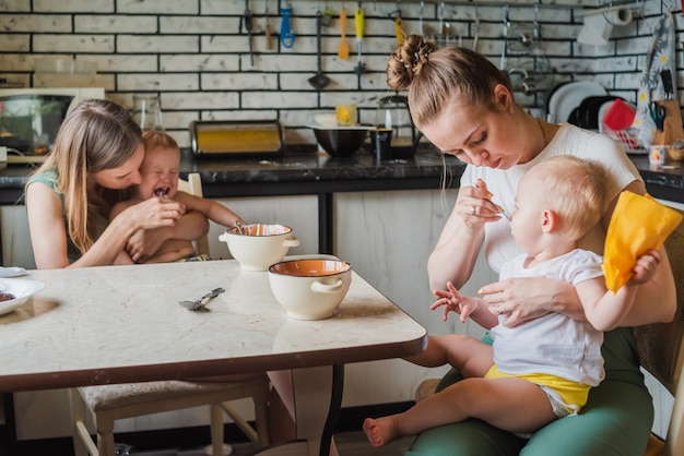 Due giovani madri insieme nutrono i loro bambini felici di latte porridge in cucina