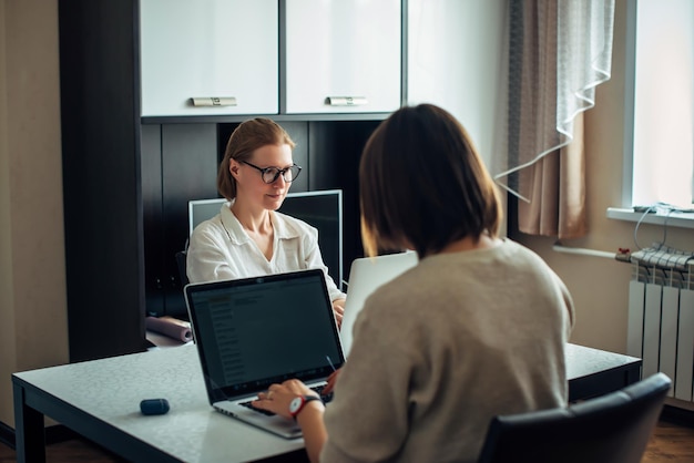 Due giovani ragazze adulte sedute a tavola una di fronte all'altra, lavorando su laptop all'interno della casa. Concentrarsi sul viso della bionda con gli occhiali. Libero professionista, coworking, lavoro a distanza, lavoro autonomo.