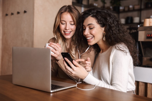 Due giovani ragazze allegre amiche sedute al tavolino del bar, divertendosi insieme, bevendo caffè, usando il laptop, ascoltando musica con gli auricolari e il telefono cellulare