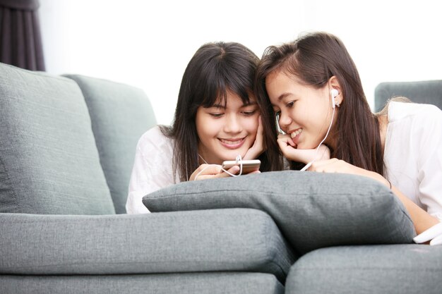 Due giovani ragazze asiatiche che guardano insieme lo smathphone mentre sono sdraiate sul divano di casa