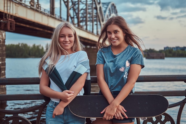 Due giovani ragazze felici hipster tengono uno skateboard e si appoggiano su un guardrail su uno sfondo del vecchio ponte.