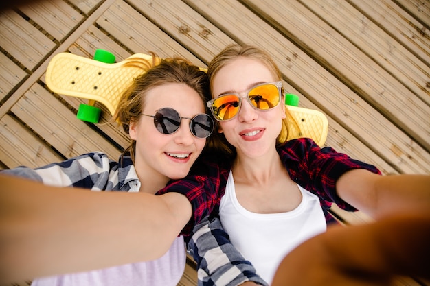 Due giovani ragazze in abito hipster che fanno selfie mentre si trovano con sul molo di legno.