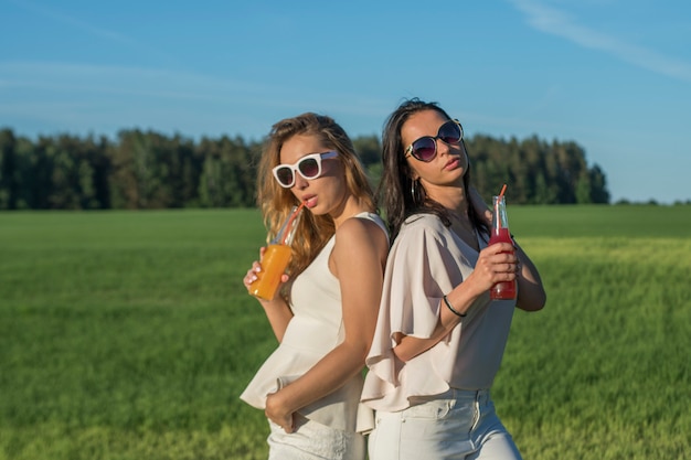 Due giovani ragazze snelle con occhiali e capelli ricci sorridono e bevono un cocktail alcolico o analcolico attraverso una cannuccia da una bottiglia in una giornata di sole estivo sorridente