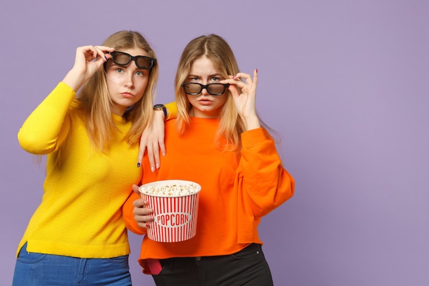 Due giovani sorelle gemelle bionde perplesse in occhiali 3d imax che guardano film in possesso di popcorn isolato su parete blu viola pastello. Concetto di stile di vita familiare di persone.