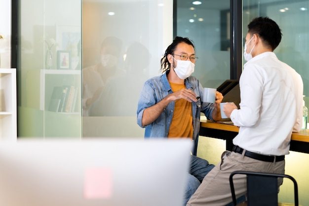 Due impiegati che parlano durante la pausa caffè nella nuova normalità con l'ufficio pratica a distanza sociale. Indossano una maschera facciale per ridurre il rischio di infezione da coronavirus COVID-19 come nuovo stile di vita normale.