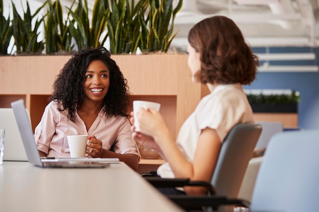 Due imprenditrici che lavorano al tavolo in un moderno ufficio a pianta aperta che bevono caffè insieme