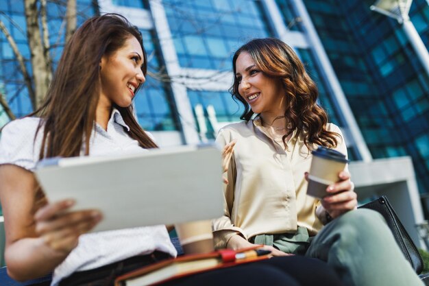 Due imprenditrici sorridenti che chiacchierano e usano la tavoletta digitale durante la pausa caffè fuori da un ufficio.