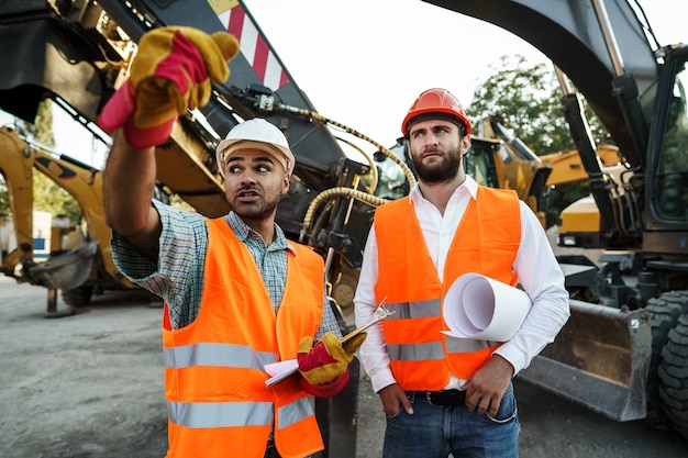 Due ingegneri discutono del loro lavoro in piedi contro le macchine edili