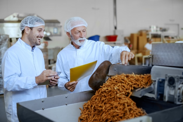 Due ispettori concentrati in divisa bianca e retine per capelli valutano la qualità del cibo. Entrambi indossano uniformi bianche e hanno retine per capelli. Interno della pianta alimentare.