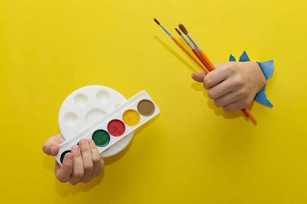 Due mani di bambini tese attraverso un foglio strappato di carta gialla tengono pennelli e colori per disegnare