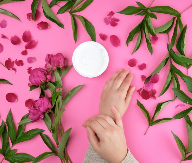 Due mani di una giovane ragazza con la pelle liscia e un vaso con crema densa e peonie in fiore bordeaux