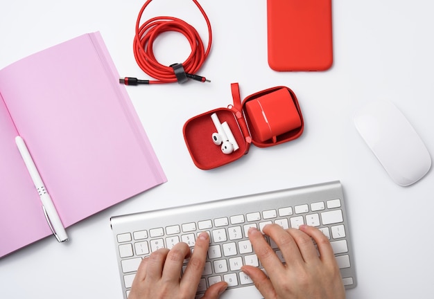 Due mani femminili e una tastiera bianca, lavoro libero professionista con un taccuino aperto, cuffie, smartphone rosso, tavolo bianco, vista dall'alto
