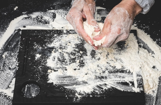 Due mani femminili impastano la pasta dalla farina di frumento bianca su una vista da tavolo nera