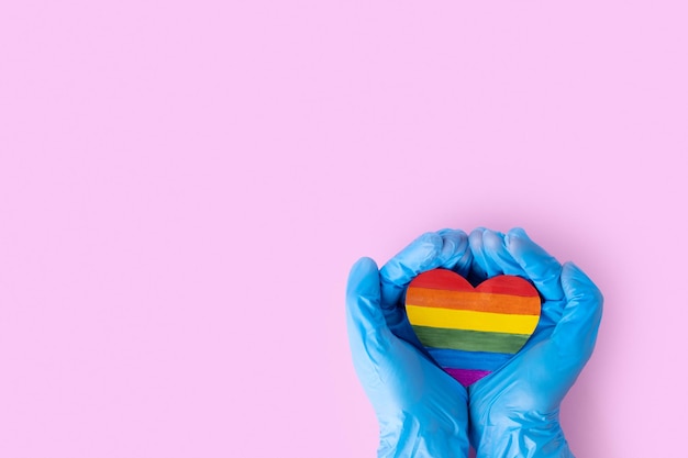 Due mani in guanti blu protettivi tengono un cuore di carta arcobaleno su uno spazio di copia di sfondo rosa Concetto LGBTQL