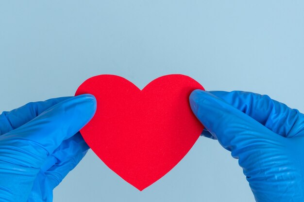 Due mani in guanti medicali blu che tengono un modello a forma di cuore rosso su sfondo blu