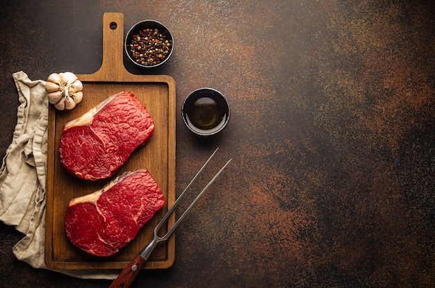 Due materie bistecche di manzo non cotte su tagliere di legno con spezie, olio d'oliva, aglio giacevano su sfondo di cemento marrone scuro con spazio di copia per il testo