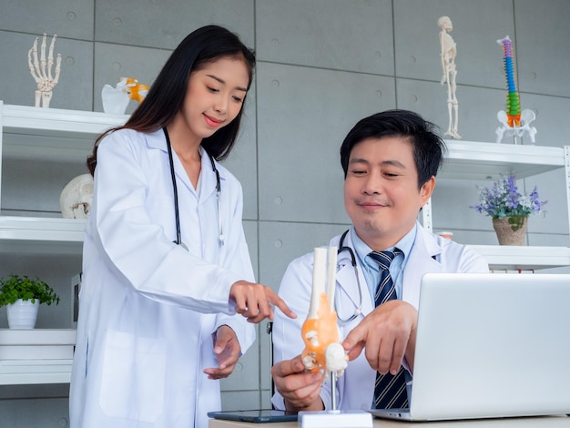 Due medici asiatici in uniforme bianca parlano e lavorano insieme nello studio medico Il medico maschio adulto consiglia alla giovane dottoressa i legamenti attorno al modello dell'osso della caviglia sulla scrivania