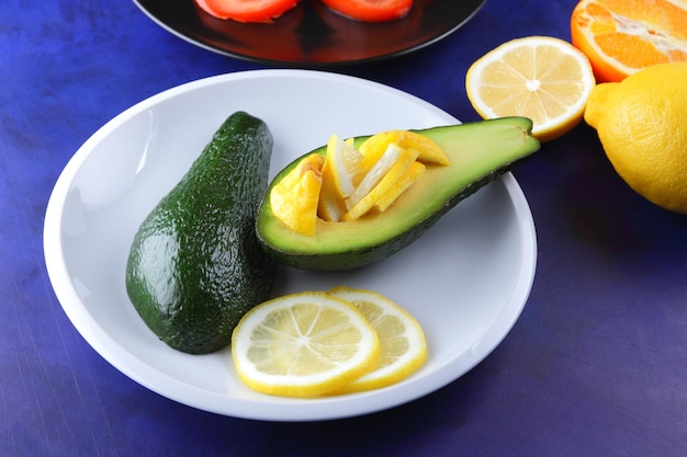 Due metà di un avocado maturo con seme su un piatto Primo piano di frutta verde e fette di agrumi e pomodoro su sfondo blu Concetto di cibo vegetariano