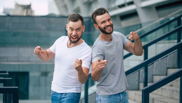 Due migliori amici barbuti moderni eccitati felici in abbigliamento casual divertendosi e indicando insieme sulla macchina fotografica all'aperto.