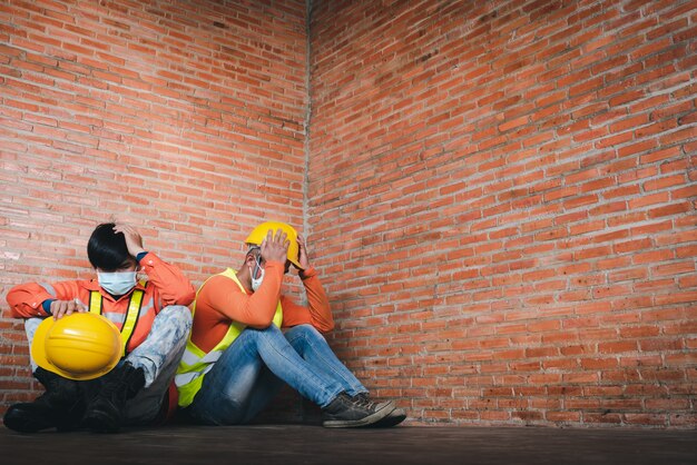 Due operai edili si pentono di essere tristi seduti sul posto di lavoro Indossare una maschera medica per prevenire Covid-19 è la disoccupazione e la crisi economica. Disoccupazione non riuscita durante Covid-19