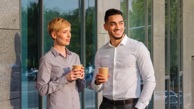 Due partner commerciali in piedi all'aperto vicino all'edificio aziendale bevono caffè parlando durante la pausa