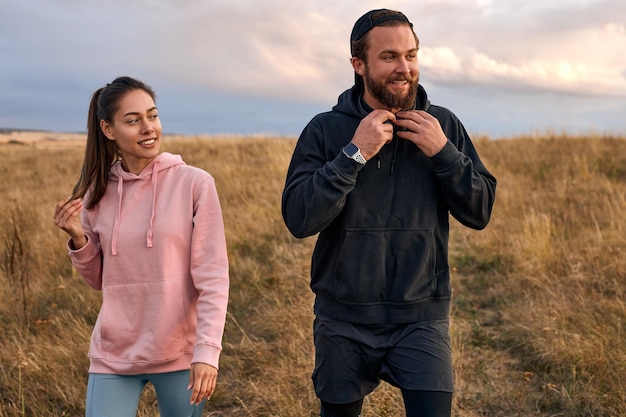 Due persone in forma felice che si preparano all'allenamento sportivo all'aperto, sul campo. bella donna e ragazzo barbuto in felpa con cappuccio godono di uno stile di vita sano. nella stagione estiva, al mattino