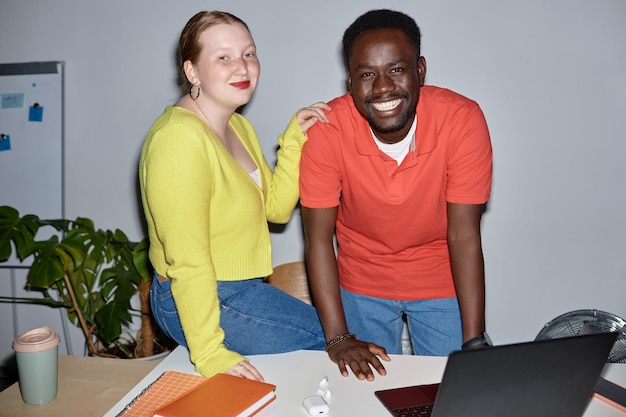 Due persone sorridenti che guardano la fotocamera sul posto di lavoro e si appoggiano alla scrivania