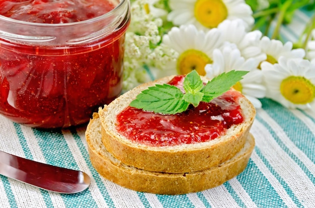 Due pezzi di pane con marmellata di fragole, un vasetto di marmellata, un coltello e un bouquet di camomilla su un tovagliolo a righe verdi