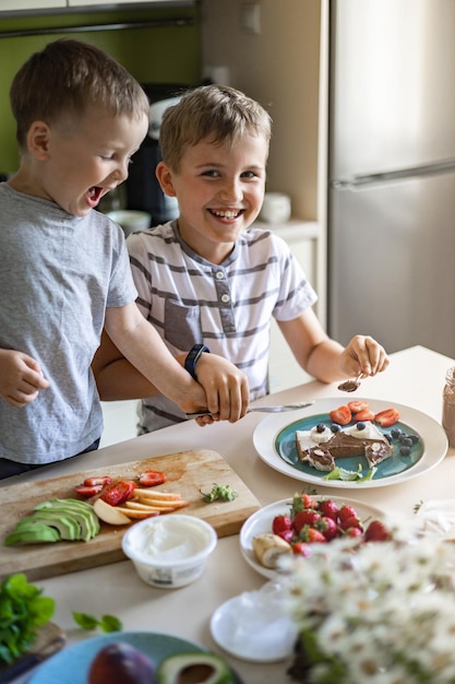 Due piccoli amici maschi che decorano il panino mattutino con frutti di bosco e pasta di cioccolato