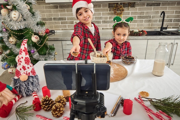 Due piccoli chef blogger, adorabili bambini caucasici, che registrano un video blog mentre cucinano insieme, mostrando l'uvetta in un cucchiaio di legno nelle loro mani a una fotocamera dello smartphone montata su un treppiede.