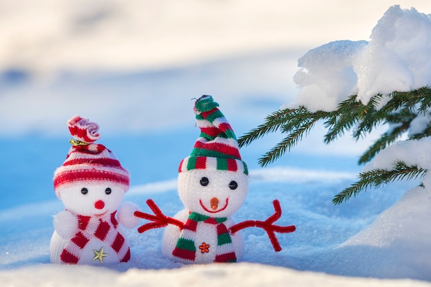 Due piccoli giocattoli divertenti baby pupazzi di neve in cappelli lavorati a maglia e sciarpe nella neve profonda all'aperto vicino al ramo di un albero di pino