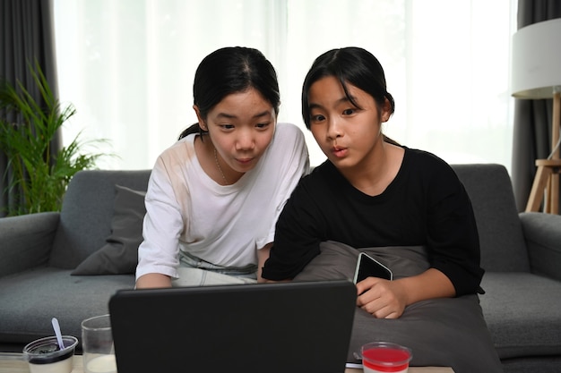 Due ragazze asiatiche che si siedono sul sofà e che guardano i cartoni animati in linea con la compressa digitale a casa