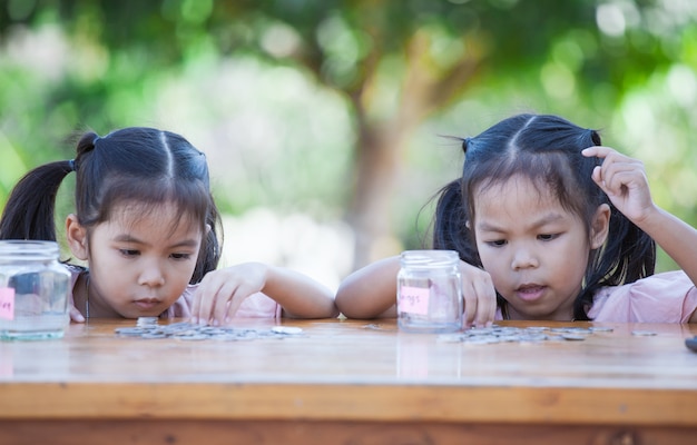 Due ragazze asiatiche del piccolo bambino che contano e che mettono insieme le monete nella bottiglia di vetro