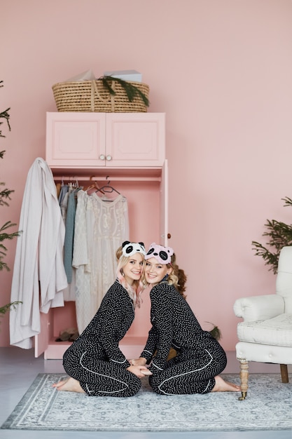 Due ragazze bionde belle, felici e sorridenti in pigiama alla moda e in eleganti maschere da notte a forma di panda siedono sul pavimento e posano all'interno