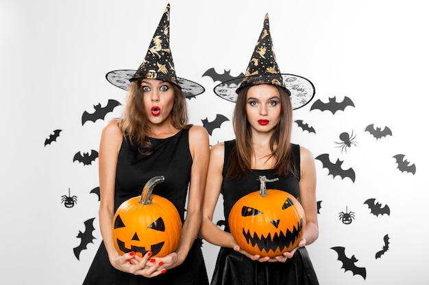 Due ragazze brune in abiti neri e cappelli da strega tengono zucche di halloween horror sullo sfondo con pipistrelli.