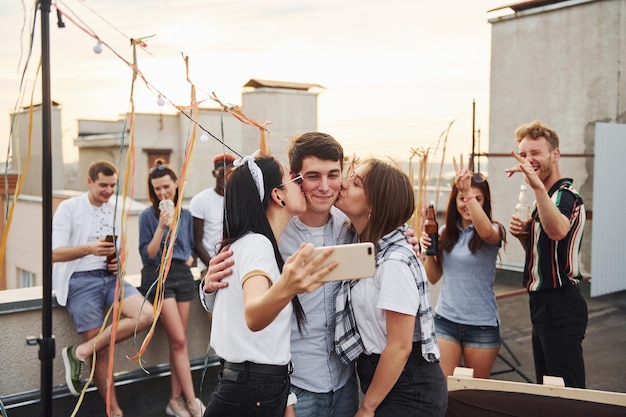 Due ragazze che baciano un uomo Prendendo selfie Un gruppo di giovani in abiti casual organizzano una festa sul tetto insieme durante il giorno