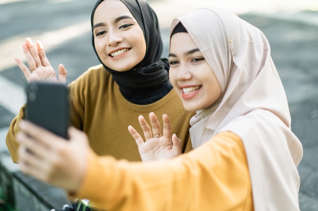 Due ragazze che indossano l'hijab con il gesto delle mani quando fanno una videochiamata insieme allo smartphone nel pomeriggio nel parco