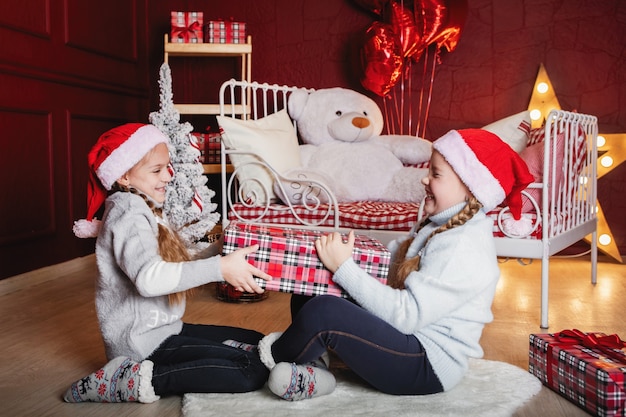 Due ragazze con le trecce in berretti rossi di Natale con scatole regalo siedono sul pavimento vicino al letto.