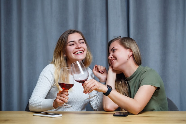 Due ragazze di aspetto europeo con i capelli biondi sono sedute al tavolo, bevono vino e ridono, si rilassano a casa, alcol