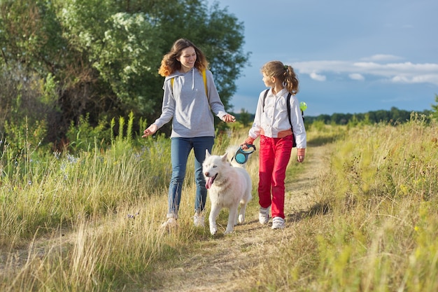 Due ragazze felici che camminano con il cane, sorelle con animali husky bianco sul prato estivo