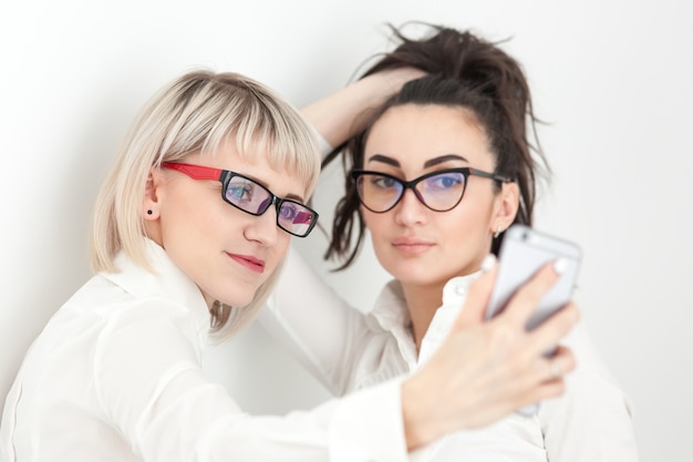 Due ragazze in camicie bianche e occhiali fanno selfie al telefono