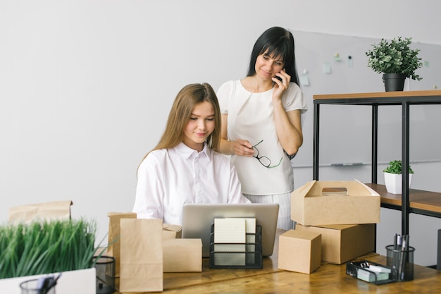 Due ragazze in ufficio stanno lavorando con un laptop e un telefono accanto a pacchetti ecologici in piedi