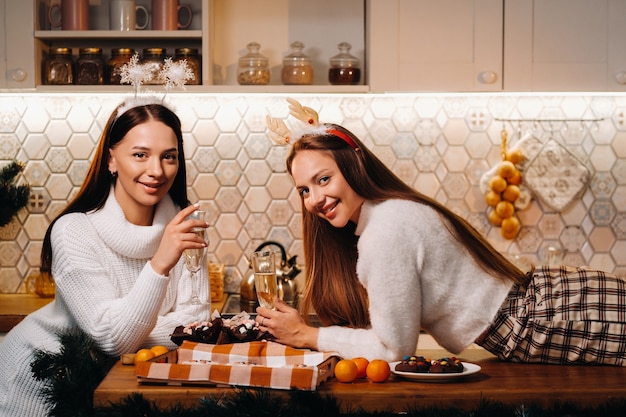 Due ragazze in un accogliente ambiente domestico con champagne in mano a Natale. Le ragazze sorridenti bevono champagne in una serata festiva