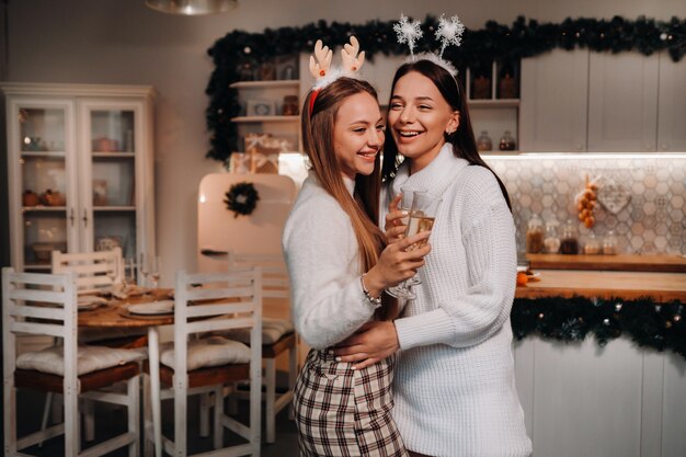 Due ragazze in un accogliente ambiente domestico con champagne in mano a Natale. Le ragazze sorridenti bevono champagne in una serata festiva