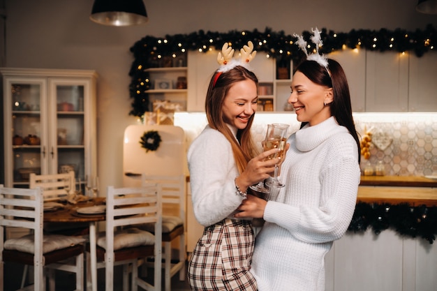 Due ragazze in un accogliente ambiente domestico con champagne in mano a Natale. Le ragazze sorridenti bevono champagne in una serata festiva