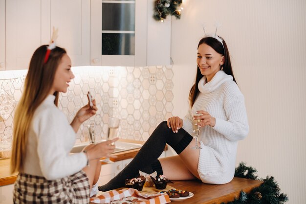 Due ragazze in un accogliente ambiente domestico in cucina con champagne in mano per Natale. Le ragazze sorridenti bevono champagne in una serata festiva