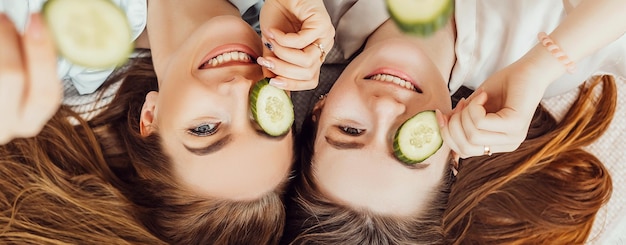 Due ragazze realizzano maschere di bellezza per viso e capelli fatte in casa Cetrioli per la freschezza della pelle intorno agli occhi Le donne si prendono cura della pelle giovane Le amiche ridono a casa sdraiate per terra sui cuscini